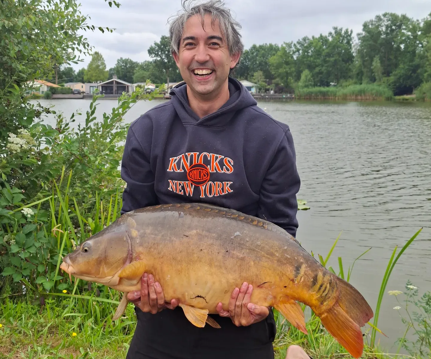 Jeroen Leidelmeijer kneeling by a lake, holding a 40 lb (18.3 kg) mirror carp across his body: a deep golden-bronze fish with a row of large scales along its back and an otherwise bare side