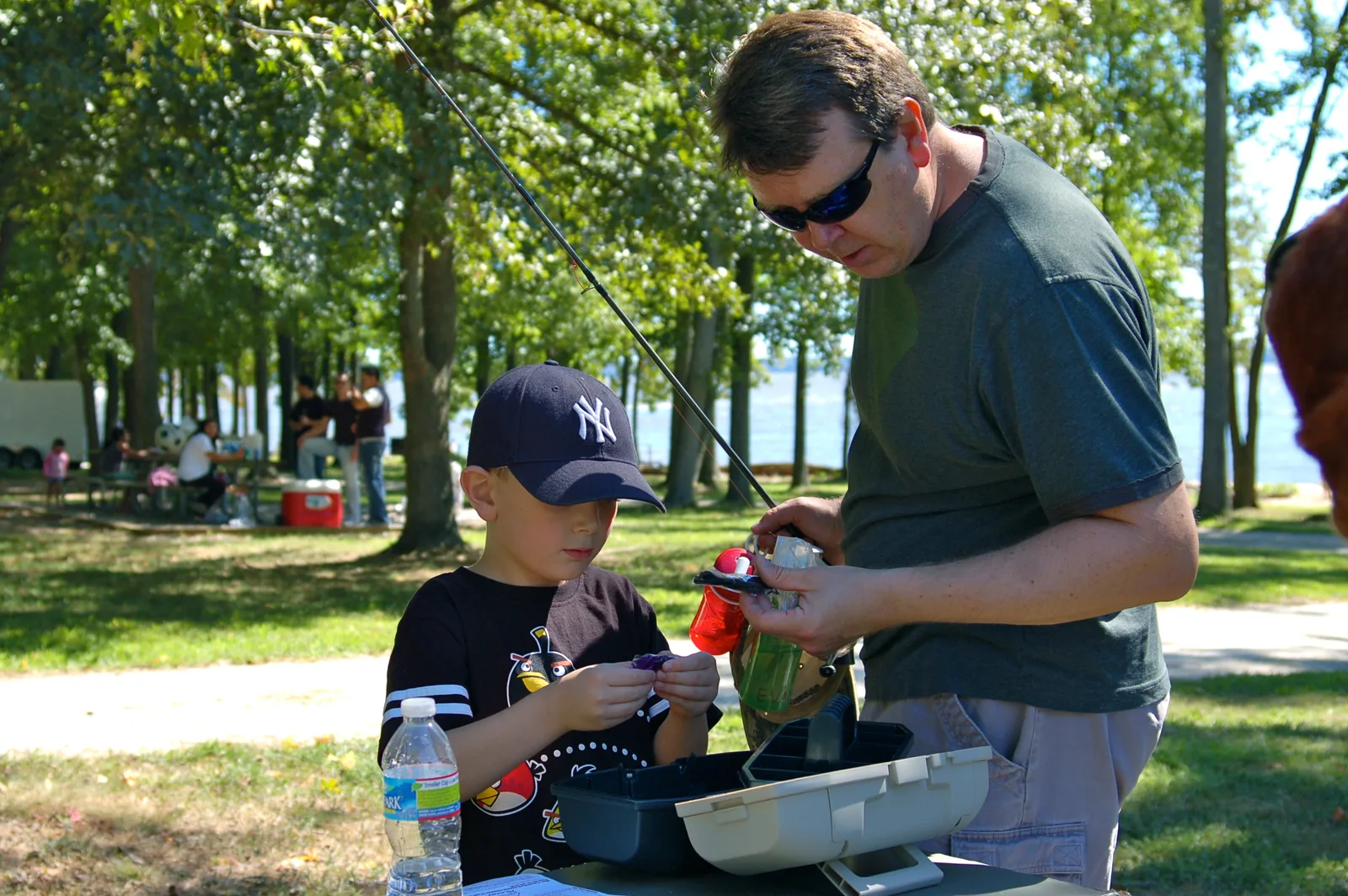 A man shows a boy the one-tray tackle box given to every child at a C.A.S.T. for Kids event, at a picnic table by the water, a fishing rod behind them