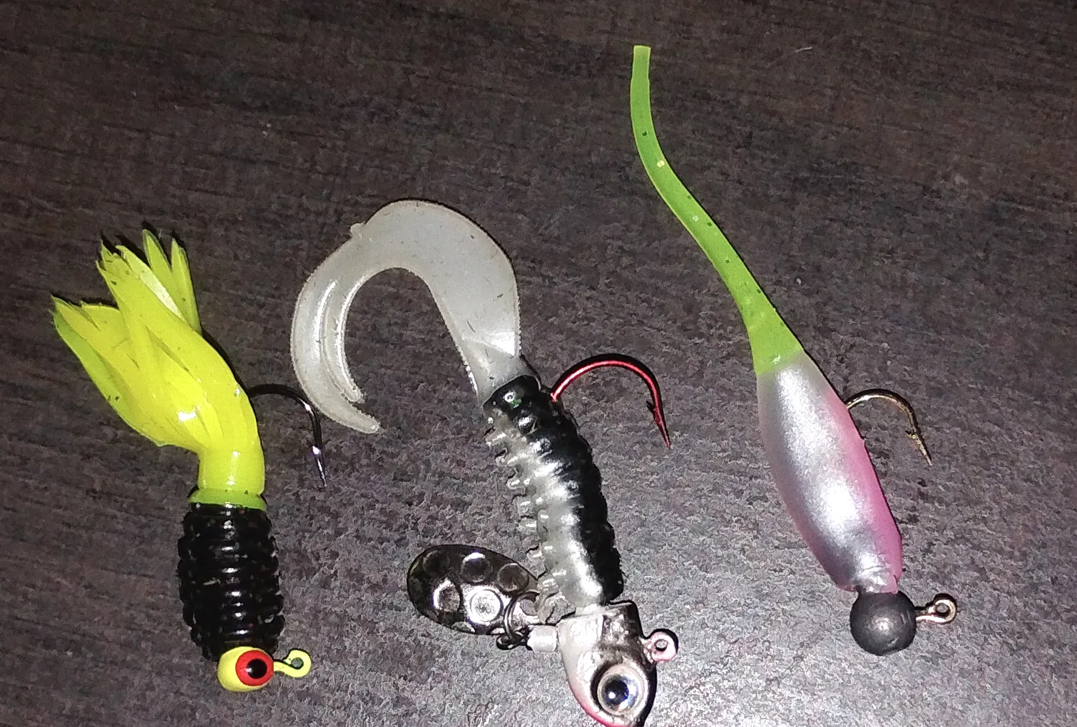 Three crappie jigs on a dark table: a chartreuse-and-black tube on a chartreuse round head with a painted eye, a white curly-tail grub on a spinner head with a small silver blade, and a pink-and-silver shad body with a thin chartreuse tail on a plain lead ball head