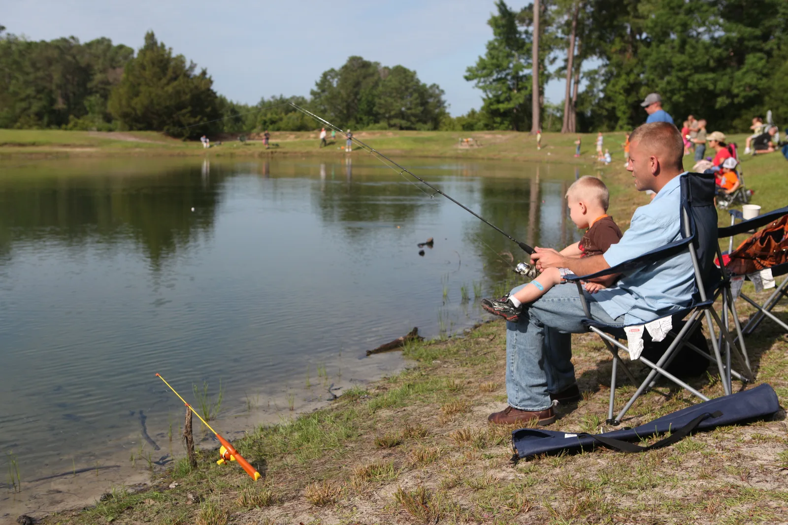 A father and his young son fishing a pond from the bank with a spinning rod and reel combo, a small child's push-button combo lying in the grass beside them