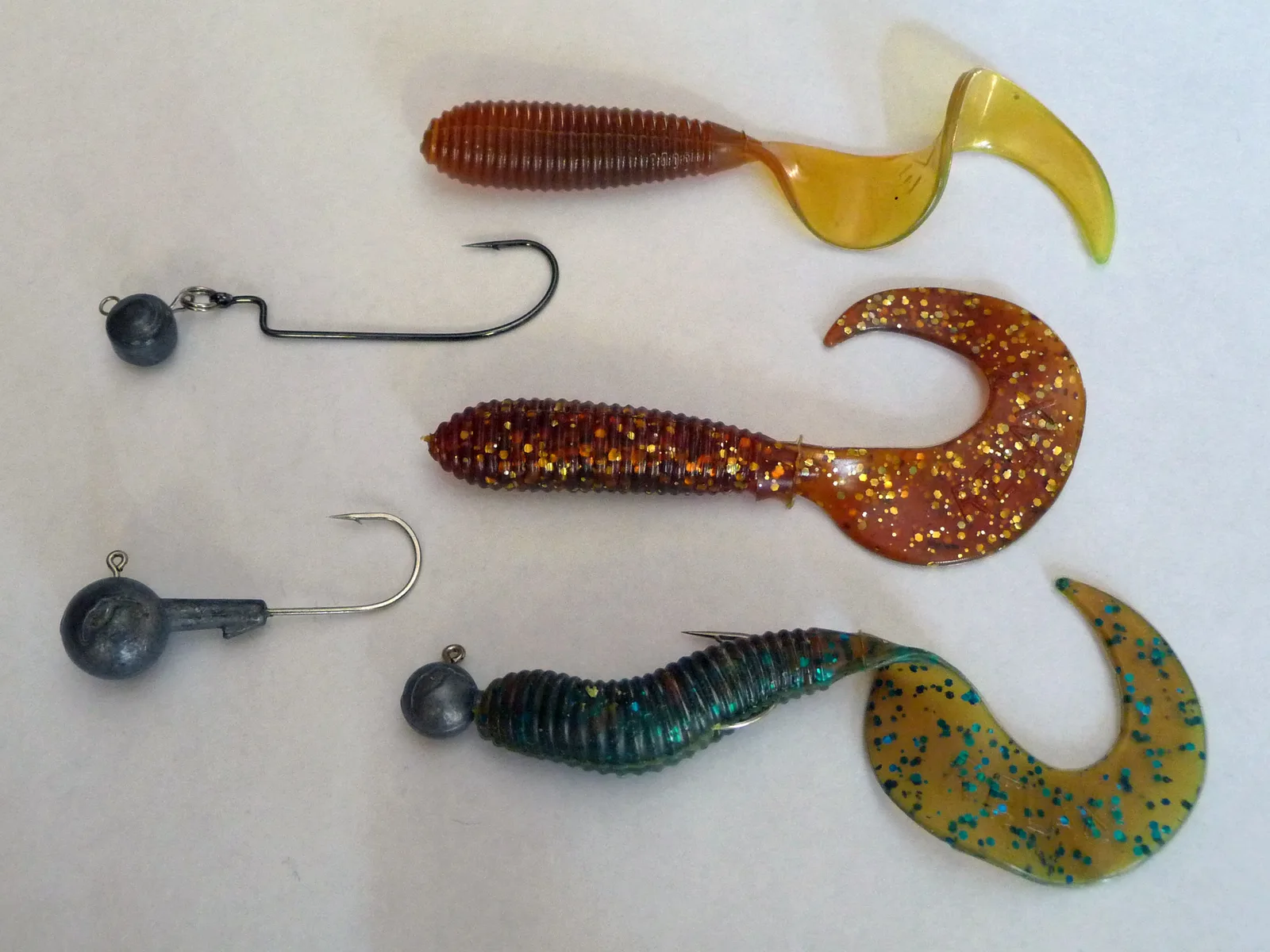 Two bare ball jig heads, one with an offset worm hook, beside three soft-plastic curly-tail grubs, one of them threaded onto a jig head
