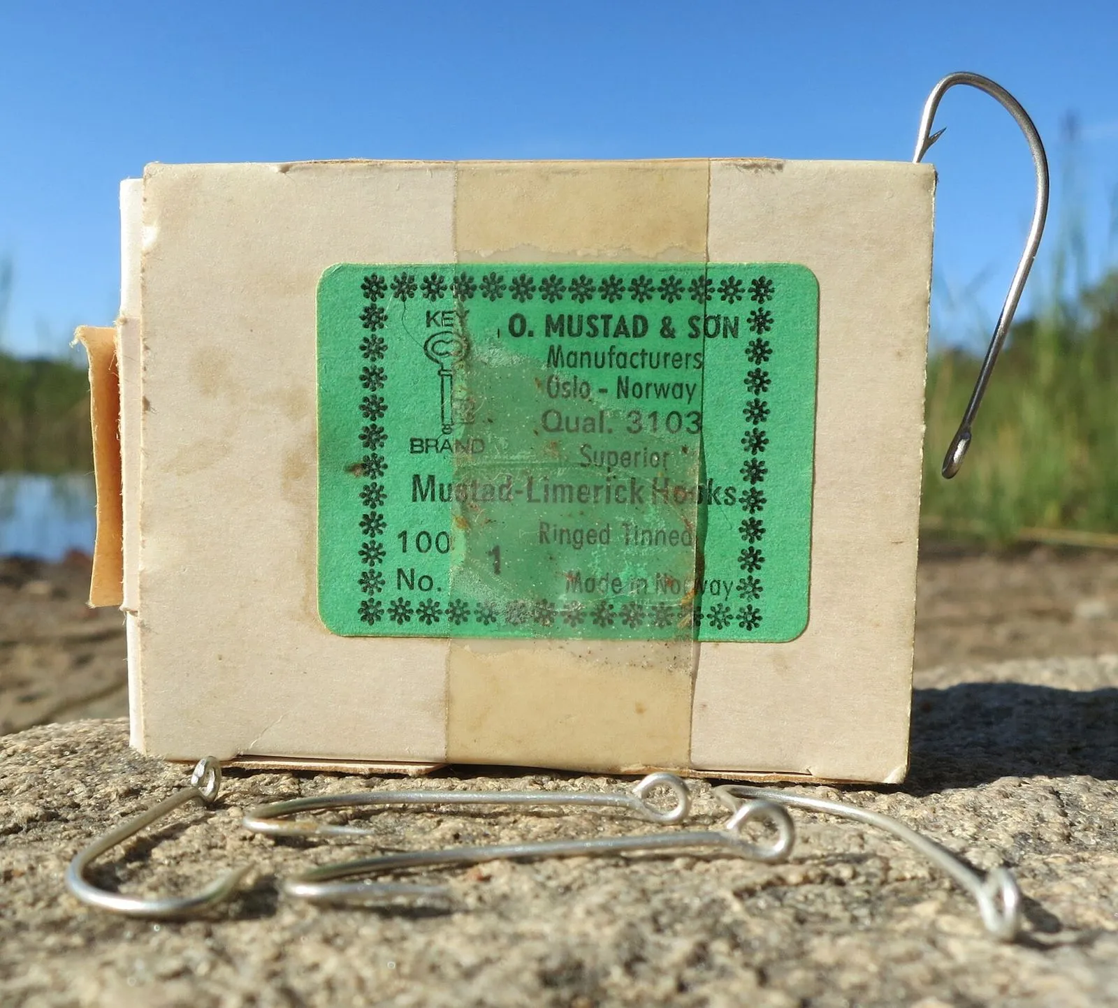 A 1970s box of Mustad fish hooks lying on sand, hooks beside it
