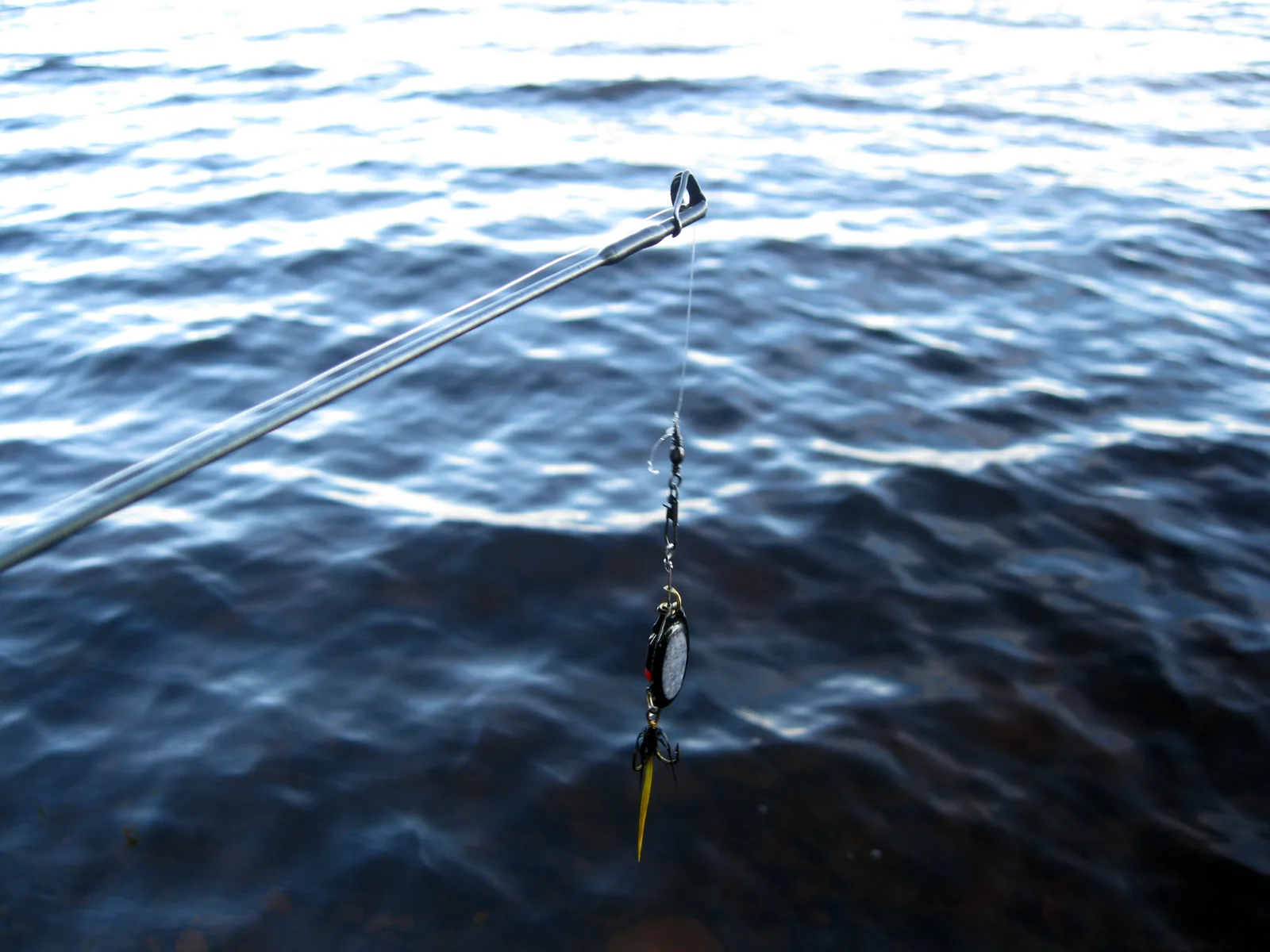 An inline spinner hanging from the tip of a spinning rod over dark lake water, a swivel tied into the line a short way above the lure
