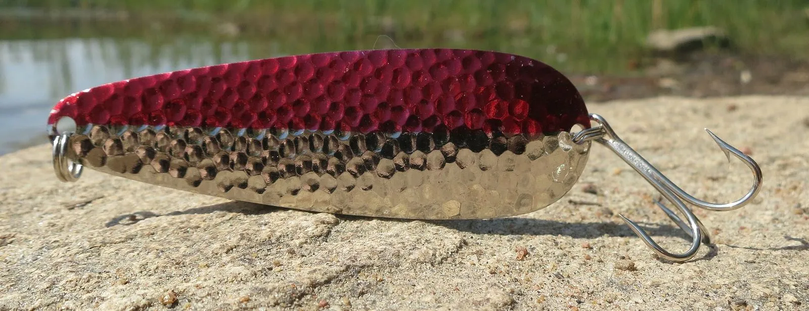 A hammered metal spoon lure with a red scale pattern and a single hook, lying on sand